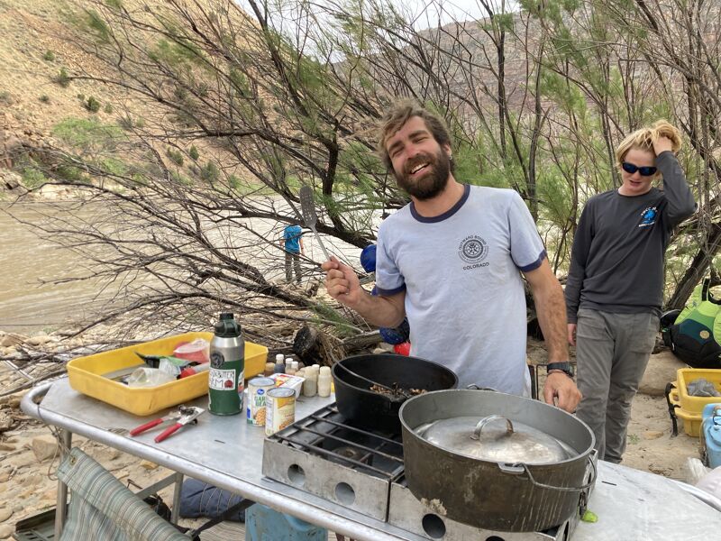 A man stands outdoors near cooking equipment, holding a utensil. He is smiling and wearing a t-shirt. Behind him, another person stands wearing sunglasses and a sweatshirt. The setting appears to be a campsite near a river or body of water, with trees and a hillside in the background. Cooking pots and other camping gear are visible on a table.